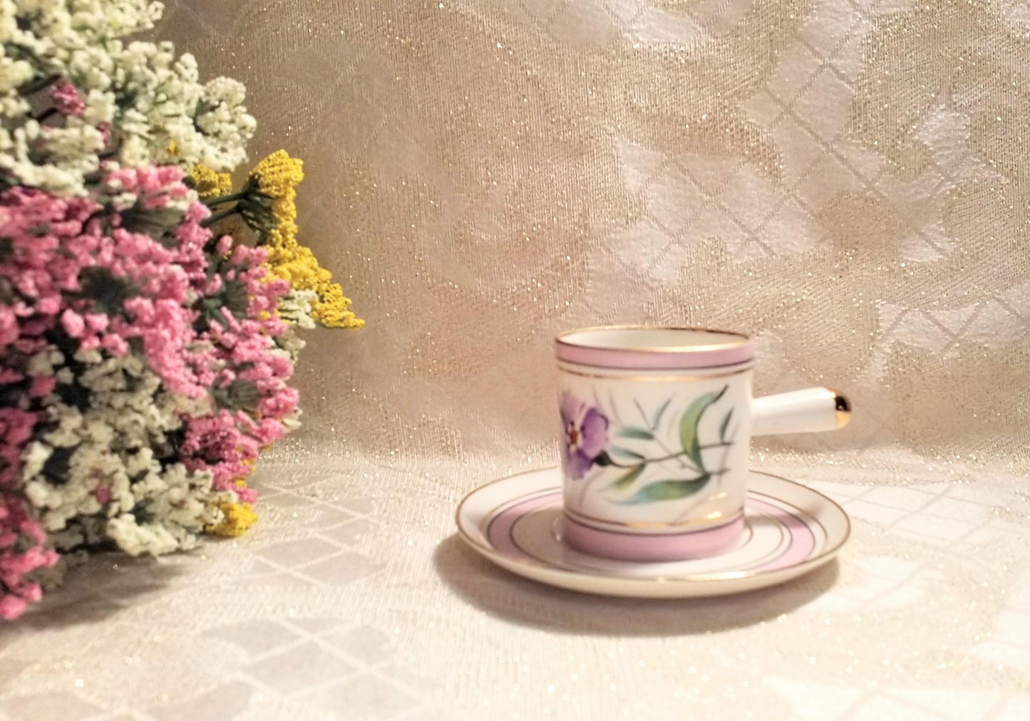 May include: A white porcelain teacup and saucer with a pink rim and gold trim. The teacup has a floral design with purple flowers and green leaves. The saucer has a pink rim and gold trim.