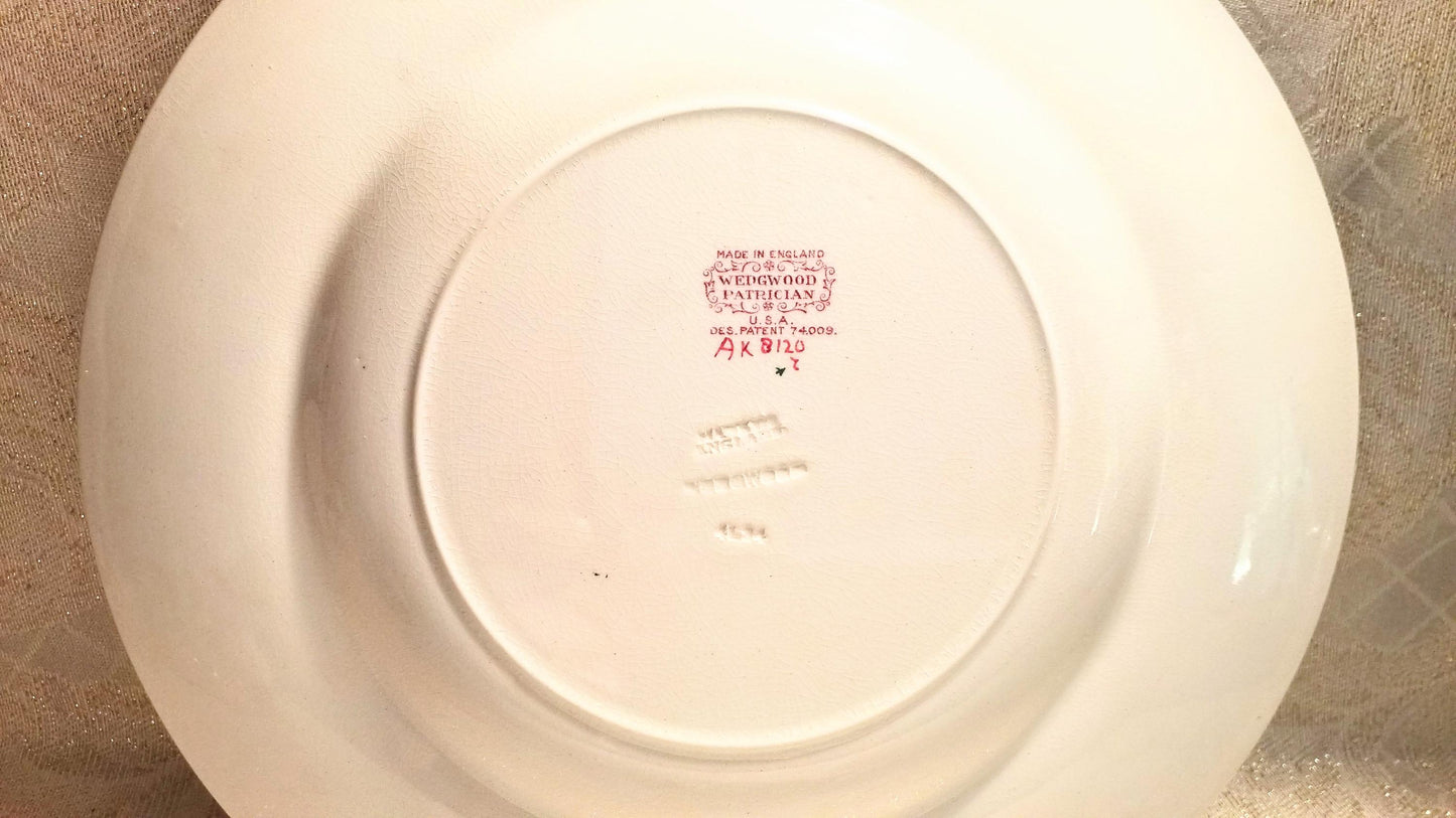 May include: A white ceramic plate with a slightly curved rim. The underside features a central circular indentation with red text and a decorative emblem. The text includes "MADE IN ENGLAND", "WEDGWOOD", and "PATRICIAN".