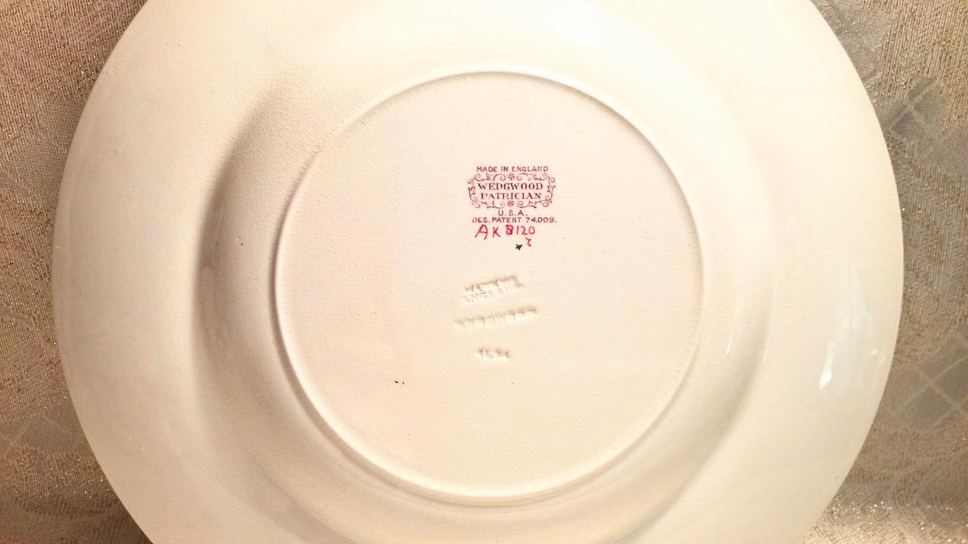 May include: A white ceramic plate with a slightly curved rim. The underside features a central circular indentation with red text and a decorative emblem. The text includes "MADE IN ENGLAND", "WEDGWOOD", and "PATRICIAN".